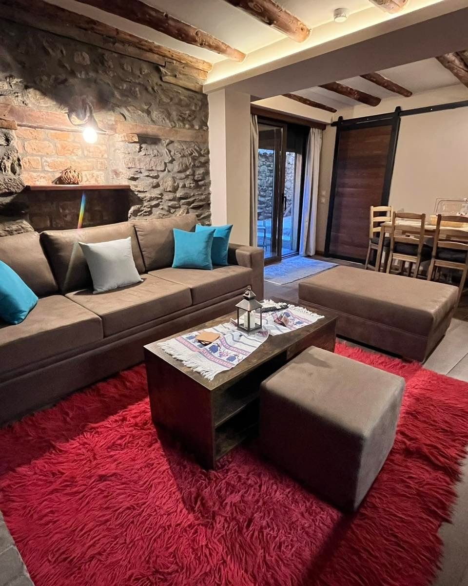 Home 2, living area with a sofa, a coffee table and a red rug, and a dining table beyond, under wooden beams.